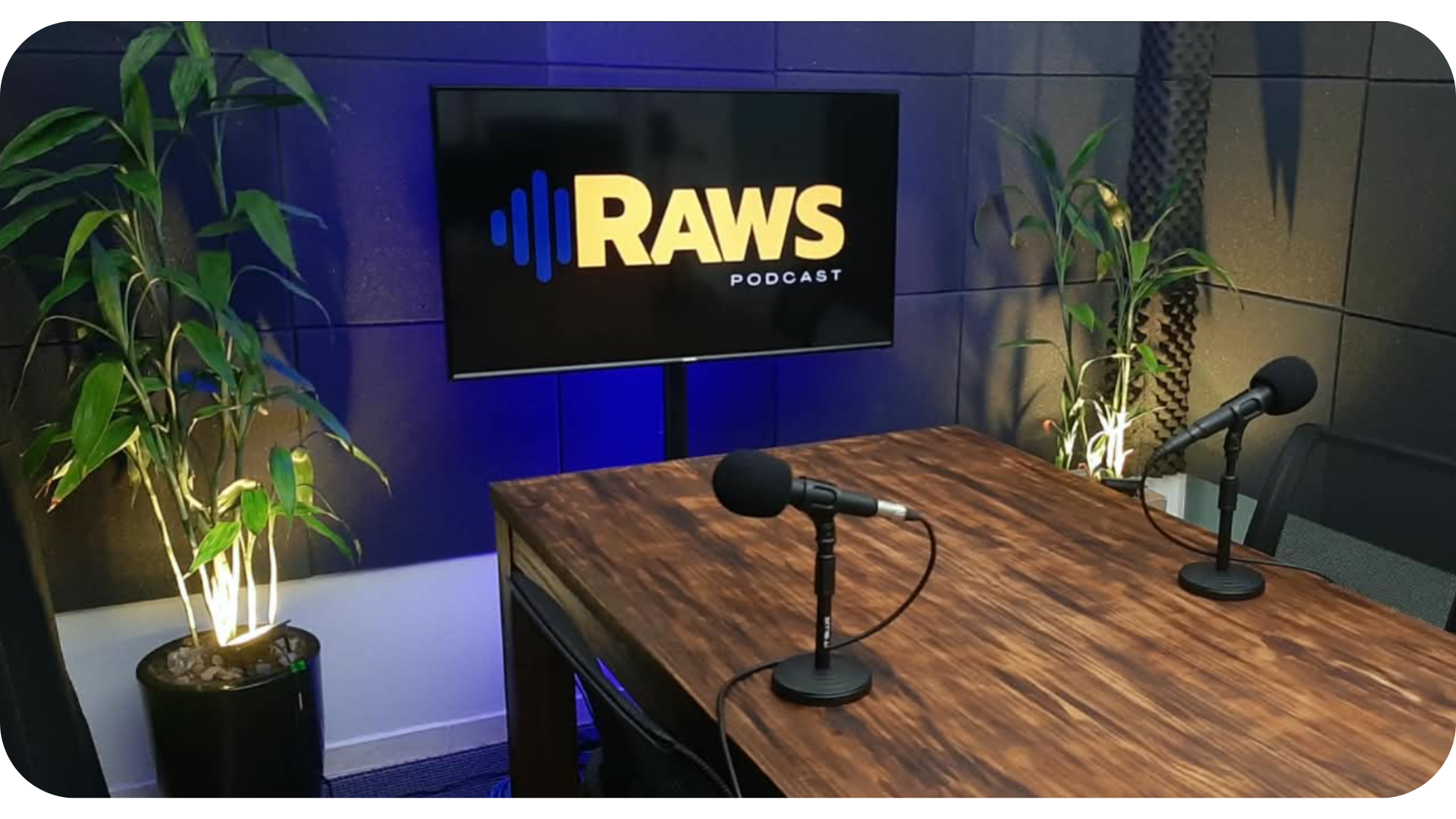 Bancada do estúdio RAWS Podcast, com microfones e a logo RAWS na TV ao fundo